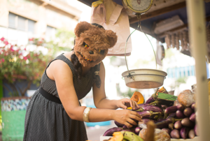 Woman wearing polka dot sleeve less dress and a brown evil bear mask, holding an eggplant posing looking at the camera standing in front of a farmers market vegetable spot
