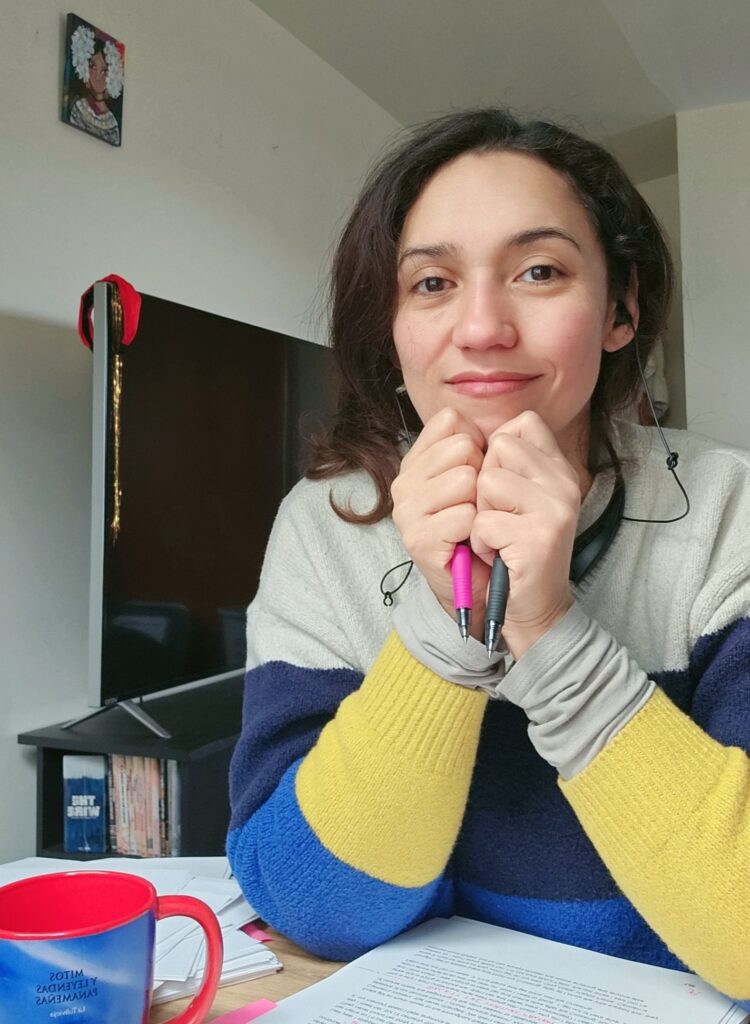 Hispanic woman smiling, hands together holding a pen, wearing a white, blue and yellow sweater. Sitting on a desk in a bedroom.