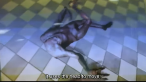 Close-up of the AI-generated humanoid creature crawling on its head across a checkered floor. Subtitle reads: “It uses the head to move.”